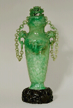 An impressive double chained celadon green jadeite vase and cover.  With dragon's head handles suspending loose rings to which each chain having ten loose double links are attatched to the cover.  Well hollowed, and splashes of emerald green well used to carve dragons in high relief.  Chinese, 1880 - 1920.  Dimensions: 23.5cm high x 10.5 wide x 8.5cm depth.