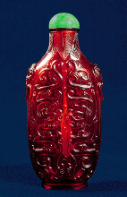 Glass, translucent ruby-red exquisitely carved with writhing and confronting archaic dragons, the neck surrounded with palm fronds, the shoulders carved with mask ring handles; the bottle of elegant, tall flattened form with a raised oval foot rim.  Chinese, Qianlong period, 1736-1795.  Stopper: Green jadeite.  Provenance: Hugh Moss Ltd, London, 1975.  Height: 8.5cm.  The design, manufacture, essence and inspiration for this beautiful glass work of art are pure Imperial: even the form itself without the decoration would imply the bottle was made at the glass works in the Forbidden City, Beijing.